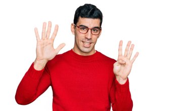 Yakışıklı İspanyol bir adam günlük kıyafetler ve gözlük takıyor. Dokuz numarayı gösteriyor ve işaret ediyor. Kendinden emin ve mutlu gülümsüyor.. 