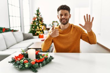 Arap genç adam noel ağacının yanında oturmuş 6 numaralı parmaklarıyla işaret ederken kendinden emin ve mutlu bir şekilde gülümsüyor.. 