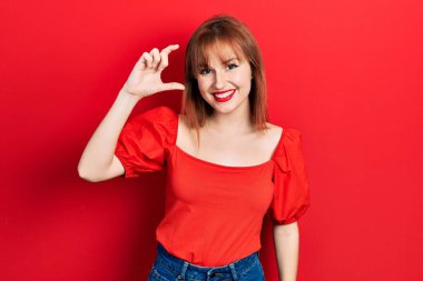 Kızıl saçlı genç kadın, günlük kırmızı tişört giyiyor gülümsüyor ve kendine güvenen el hareketleriyle küçük beden işareti yapıyor, parmaklarıyla bakıyor ve kameraya bakıyor. ölçüm kavramı. 