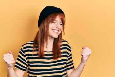 Kızıl saçlı, yün şapka takan genç bir kadın çok mutlu ve heyecanlı, kolları havada, gülümseyen ve başarı için bağıran, kazanan bir jest yapıyor. kutlama konsepti. 