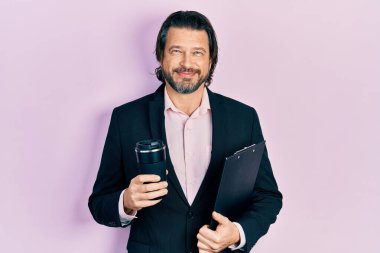 Orta yaşlı beyaz bir adam iş kıyafetleri içinde bir fincan kahve içerken yüzünde mutlu ve havalı bir gülümsemeyle gülümsüyor. diş gösterme. 