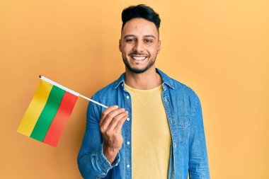 Elinde Litvanya bayrağı tutan genç bir Arap adam pozitif ve mutlu görünüyor ve kendine güvenen bir gülümsemeyle dişlerini gösteriyor. 