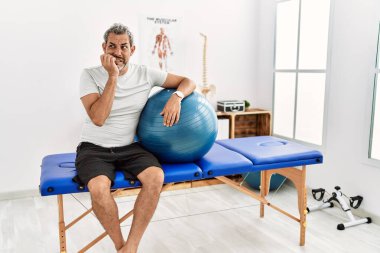 Orta yaşlı İspanyol bir adam, ağrı kesici kliniğinde elinde pilates topu tutarken gergin ve eli ağızda, tırnaklarını yiyormuş gibi görünüyor. anksiyete. 