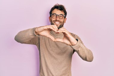 Balıkçı yaka kazak ve gözlük takan sakallı yakışıklı aşık bir adam elleriyle kalp sembolü yapıyor. romantik kavram. 