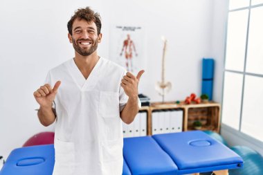 Ağrı kesici kliniğinde çalışan genç, yakışıklı fizyoterapist. Arka tarafı işaret ediyor. El ve baş parmakları havada, kendinden emin. 