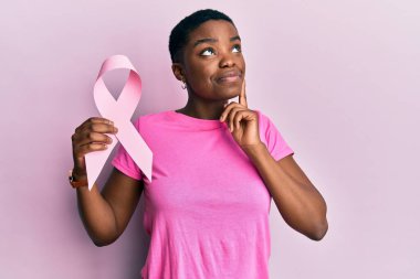 Genç Afro-Amerikalı kadın pembe kanser kurdelesi takmış ciddi bir yüz ifadesiyle eli çenesinde soruyu düşünüyor, kafa karıştırıcı bir fikir. 
