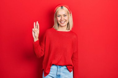 Güzel sarışın kadın gündelik kırmızı süveter giyer ve üç numaralı parmaklarıyla gülümseyerek mutlu ve kendinden emin bir şekilde gösterir.. 
