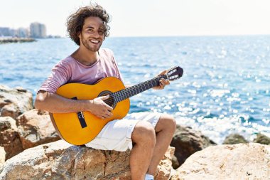 Genç İspanyol adam klasik gitar çalıyor plajda rock 'ın üzerinde oturuyor..