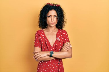 Yazlık elbise giyen genç Latin kız kuşkucu ve gergin. Yüzünde çapraz kollu onaylamayan bir ifade var. negatif kişi. 