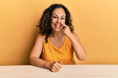 Güzel orta yaşlı kadın, gündelik giysiler içinde masaya oturmuş, parmaklarıyla yüzünü ve burnunu göstererek neşeli bir şekilde gülümsüyor. güzellik konsepti 