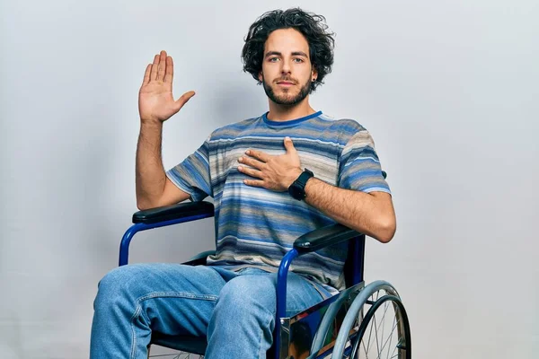 Handsome hispanic man sitting on wheelchair swearing with hand on chest and open palm, making a loyalty promise oath
