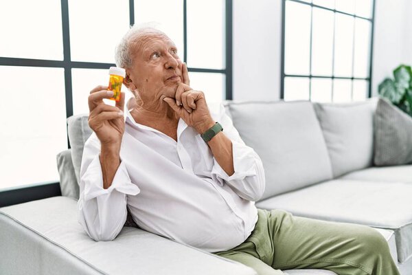 Senior man holding pills with hand on chin thinking about question, pensive expression. smiling and thoughtful face. doubt concept. 