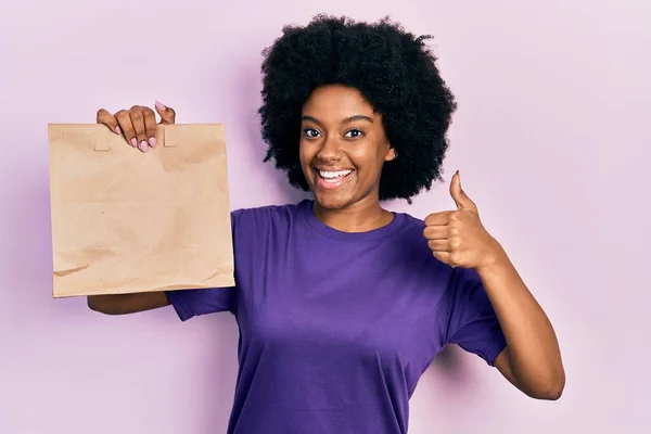 若いアフリカ系アメリカ人の女性を持って紙の袋を奪う笑顔幸せと肯定的な 親指を行う優れた承認サイン — ストック写真