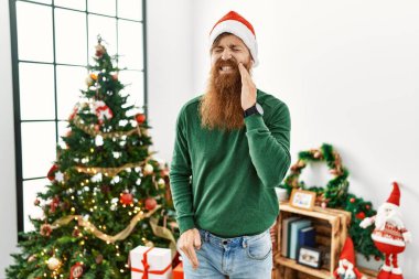 Uzun sakallı kızıl saçlı adam diş ağrısı ya da diş hastalığından dolayı Noel ağacından şapkasını takıp dudaklarına acı dolu bir ifadeyle dokunuyor. dişçi 