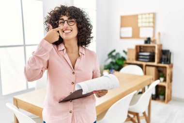 Ofiste iş elbisesi giymiş genç bir ortadoğulu kadın, el ve burunla işaret ediyor, neşeli bir şekilde gülümsüyor. güzellik konsepti 