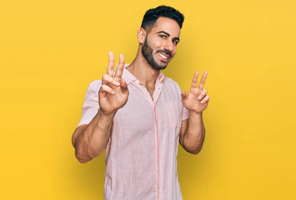 勝利のサインを行う指を示すカメラを見て笑みを浮かべてカジュアルなシャツを着て髭を持つヒスパニック系の男 — ストック写真