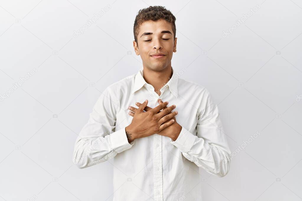 Young handsome hispanic man standing over isolated background smiling with hands on chest with closed eyes and grateful gesture on face. health concept. 
