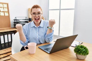 Ofiste çalışan genç kızıl saçlı kadın bilgisayarını kullanarak çok mutlu ve heyecanlı bir şekilde kollarını kaldırıp, gülümseyerek ve başarı için çığlık atarak kazanan bir jest yapıyor. kutlama konsepti. 