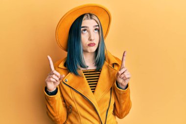 Sarı şapkalı ve deri ceketli genç bir kız üzgün ve üzgün görünüyor, parmaklarıyla yön gösteriyor, mutsuz ve depresif.. 