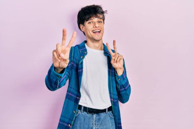 Yakışıklı hippi genç adam, günlük beyaz tişört ve klasik tişört giyip kameraya bakıp parmaklarını zafer işareti yaparken gösteriyor. İki numara.. 