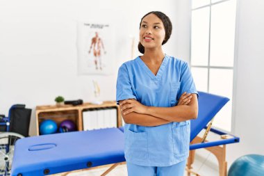 Klinikte kollarını kavuşturmuş duran fizyoterapist üniforması giyen genç Latin kadın.