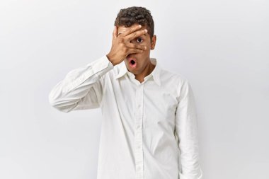 Genç, yakışıklı İspanyol bir adam izole edilmiş arka planda duruyor. Şok olmuş bir şekilde yüzünü ve gözlerini kapıyor. Utanmış bir ifadeyle parmaklarının arasından bakıyor.. 