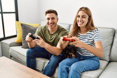 Genç İspanyol çift gülümseyerek evde köpekle kanepede oturup video oyunu oynuyor..