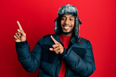 Sakallı, kışlık şapkalı, ceketli, gülümseyen ve kameraya bakan iki eliyle ve parmaklarıyla yüzünü gösteren Afrikalı genç bir Amerikalı.. 