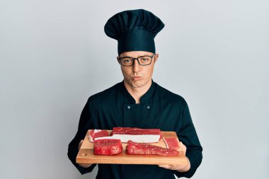 Yakışıklı genç adam, şef üniforması içinde çiğ etle pano tutuyor ve sıkıntıdan endişe ediyor. Kızgın ve korkmuş. hüzünlü ifade. 
