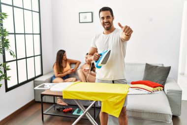 Evde giysilerini ütüleyen genç bir İspanyol, olumlu jestleri elle onaylıyor, gülümsüyor ve başarı için seviniyor. kazanan jest. 