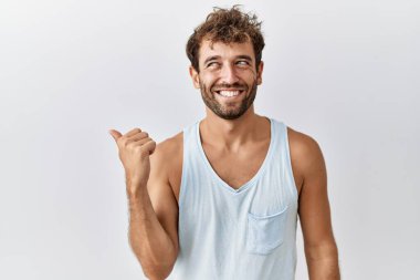 Genç yakışıklı adam izole edilmiş arka planda gülümsüyor, gülümsüyor ve başparmağıyla yan tarafa bakıyor.. 
