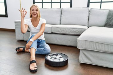 Genç beyaz kadın elektrikli süpürgeyle evde oturuyor gülümsüyor ve el ve parmaklarıyla 