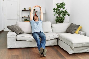 Orta yaşlı, beyaz bir adam, elleri başının üstünde, evdeki kanepede oturuyor.