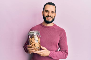 Sakallı genç adam elinde çikolata parçacıklı kurabiye kavanozuyla pozitif ve mutlu görünüyor ve kendine güvenen bir gülümsemeyle dişlerini gösteriyor 