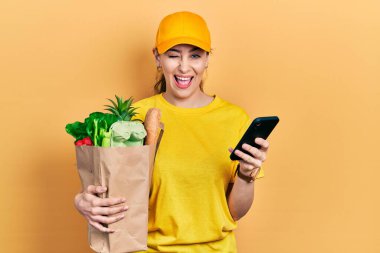 Genç İspanyol kadın elinde alışveriş torbası akıllı telefon kullanarak kameraya seksi bir ifadeyle bakıyor, neşeli ve mutlu bir yüzle.. 