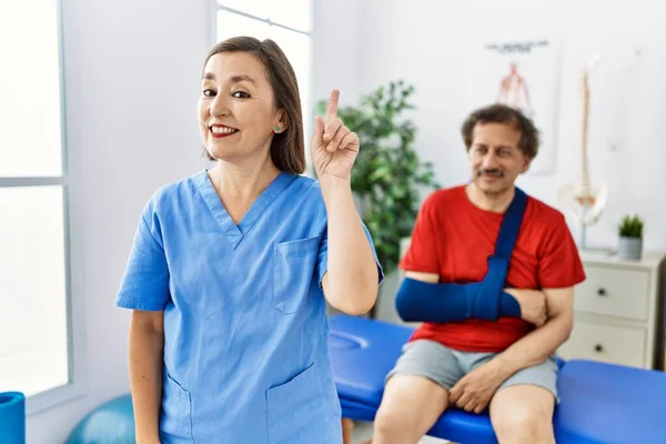 Middle age doctor woman with patient with arm injury at rehabilitation clinic smiling with an idea or question pointing finger up with happy face, number one