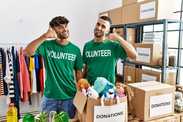 Young gay couple wearing volunteer t shirt at donations stand smiling doing phone gesture with hand and fingers like talking on the telephone. communicating concepts.