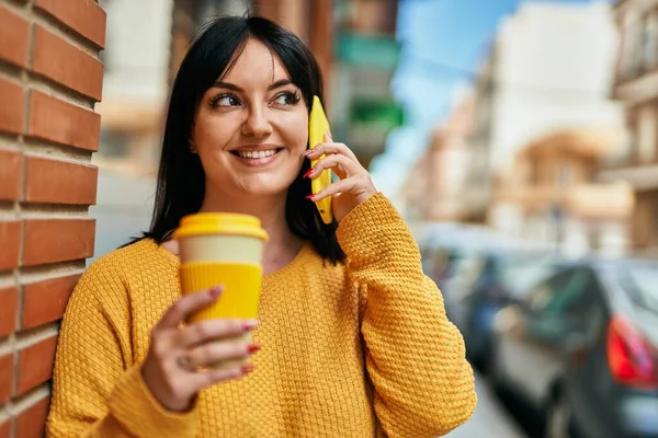 Young brunette woman speaking on the phone and drinking a cup of coffee leaning on bricks wall