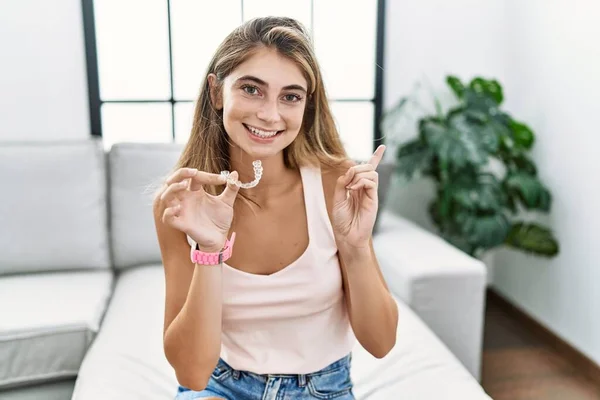 Young blonde woman holding invisible aligner orthodontic cheerful with a smile on face pointing with hand and finger up to the side with happy and natural expression