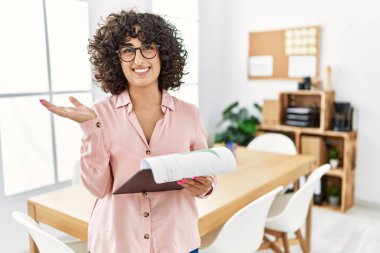 İş yerinde giyinmiş genç bir ortadoğulu kadın avuçlarını açıp fotokopi alanını gösteriyor, gülümseyerek mutlu bir gülümseme sergiliyor. 