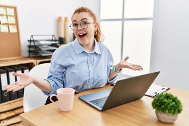 Ofiste çalışan genç kızıl saçlı kadın bilgisayar laptopu kullanıyor. Kollarını açıp gülümsüyor. Dostça karşılama, olumlu ve kendinden emin selamlar. 