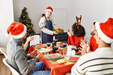 Bir grup insan el çırpıyor ve masada oturuyor. Ayakta duran ve elinde kavrulmuş hindi tutan kadın Noel 'i evde kutluyor..