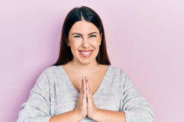 Gündelik giysiler giyen genç İspanyol kadın elleriyle dua ediyor af diliyor kendinden emin bir şekilde gülümseyerek.. 