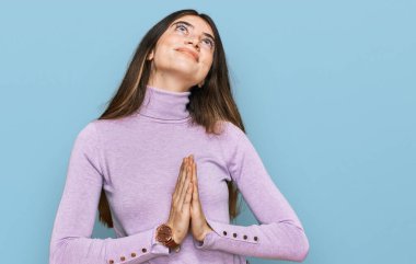 Balıkçı yaka kazak giyen genç ve güzel bir kız elleri bağlı yalvarıyor ve dua ediyor. Yüzünde umut ifadesi var. Çok duygusal ve endişeli. Yalvarıyor. 