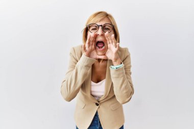 Orta yaşlı sarışın iş kadını izole edilmiş arka planda duruyor ve elleri ağzında kızgın bir şekilde bağırıyor. 