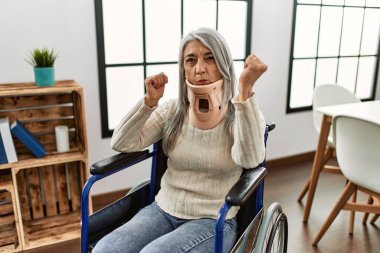 Orta yaşlı, gri saçlı, tekerlekli sandalyede oturan, tasmalı, sinirli ve sinirli bir kadın. Öfkeyle bağırıyor, öfkeyle bağırıyor ve el kaldırıyor. 