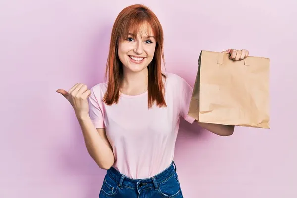 Pelirroja Joven Sosteniendo Llevar Bolsa Papel Apuntando Pulgar Hacia Lado — Foto de Stock