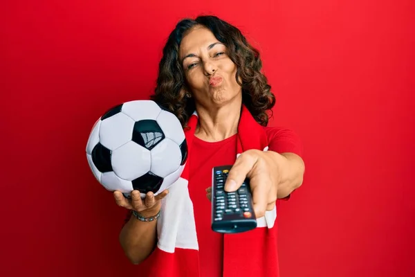 Middle age hispanic woman football hooligan holding ball using tv control looking at the camera blowing a kiss being lovely and sexy. love expression.