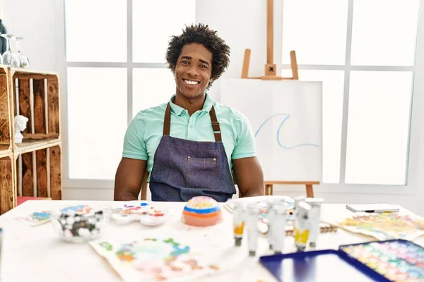 若いアフリカ系アメリカ人の男性がアートスタジオでテーブルの上に座り 前向きな笑顔で立って笑顔で歯を見せています — ストック写真