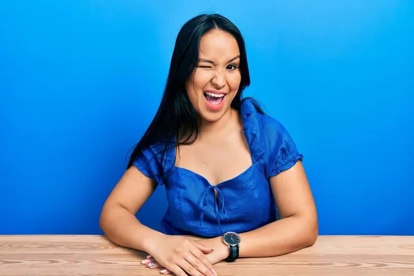 Beautiful hispanic woman with nose piercing sitting on the table winking looking at the camera with sexy expression, cheerful and happy face.